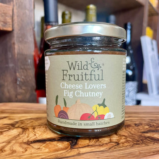 A jar of Wild & Fruitful Cheese Lovers Fig Chutney sits on a rustic wooden surface with a blurred kitchen or pantry background.